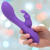 A hand with pink-painted nails holds the CalExotics Flex Bend & Hold Double Rumbler Rabbit Vibrator, featuring a curved shaft and rabbit-shaped clitoral stimulator, against a softly blurred background with colorful bokeh lights.